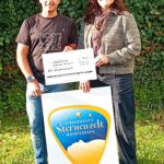 Eine Person in einem T-Shirt und eine Person in einem grauen Pullover halten einen großen Scheck vor einem Schild des Kinderhospizes Sternenzelt Mainfranken. Im Hintergrund sind grüne Pflanzen zu sehen.