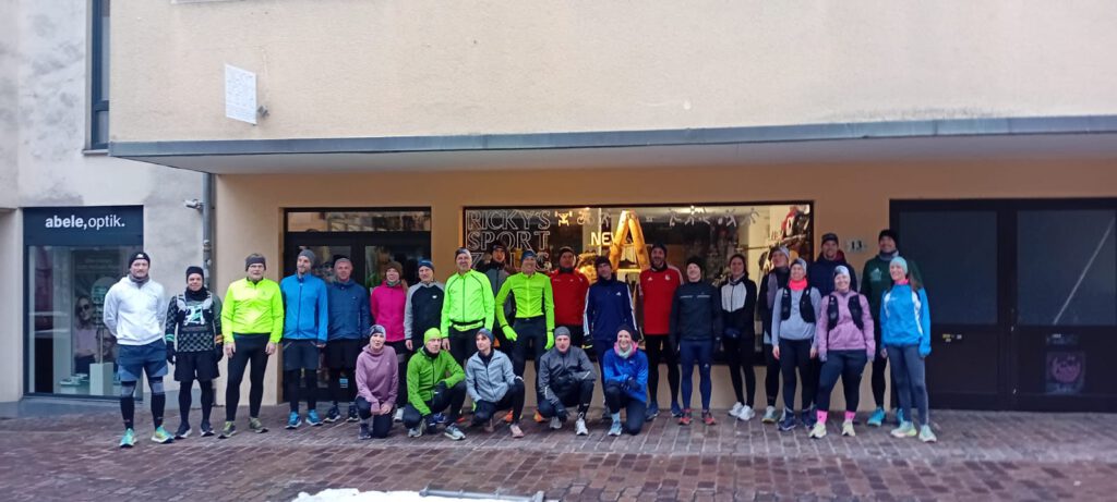 Eine Gruppe von 25 Personen in Sportkleidung steht vor einem Geschäft mit Schaufenster. Alle Teilnehmer haben verschiedene Oberteile in bunten Farben an und lächeln in die Kamera.