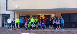 Eine Gruppe von 25 Personen in Sportkleidung steht vor einem Geschäft mit Schaufenster. Alle Teilnehmer haben verschiedene Oberteile in bunten Farben an und lächeln in die Kamera.