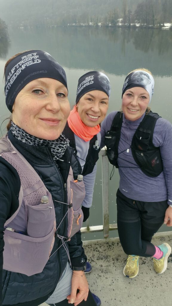Drei Frauen in Outdoor-Laufkleidung posieren auf einer Brücke mit Blick auf einen See im Hintergrund. Alle tragen Stirnbänder und Rucksäcke, bereit für eine Lauf-Tour.