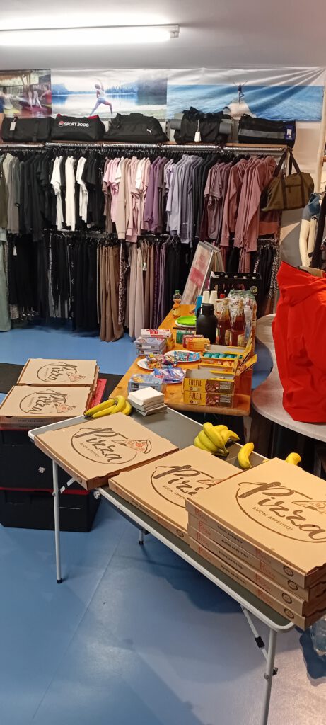 Eine Verkaufsfläche mit Regalen, die Kleidung aus Stoff zeigen. Im Vordergrund stehen Pizza-Boxen, frische Bananen und verschiedene Lebensmittel sowie Getränke auf einem Tisch.