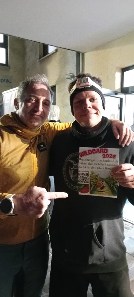 Zwei Männer posieren in einem Raum. Der Mann links trägt eine gelbe Jacke und zeigt auf ein Blatt Papier, das der Mann rechts hält. Das Blatt zeigt den Text "WILDCARD 2026". Beide lächeln und wirken erfreut.