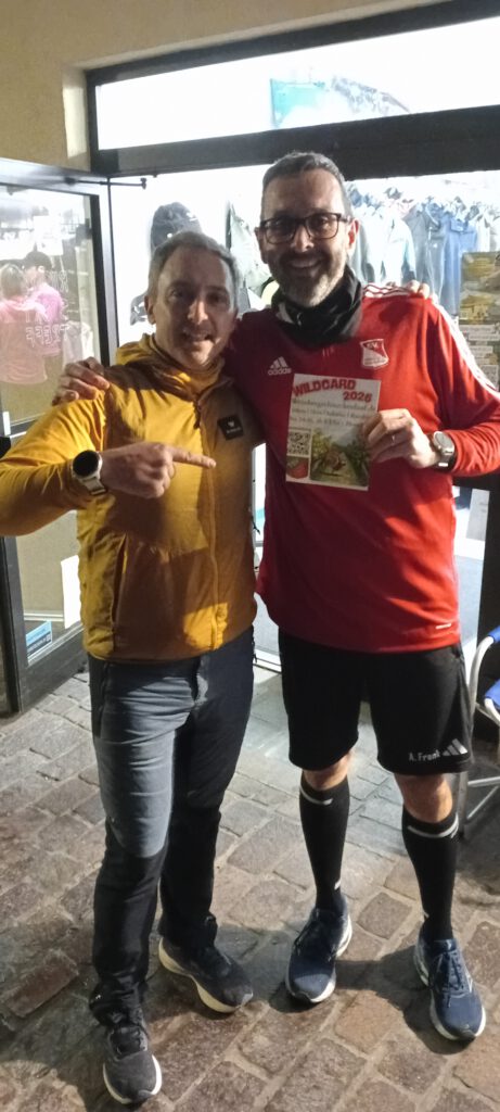 Zwei Männer posieren lächelnd vor einem Geschäft. Der Mann links trägt eine gelbe Jacke, der Mann rechts ein rotes Sporttrikot und hält ein Informationsblatt in der Hand. Beide zeigen mit dem Finger auf das Blatt.