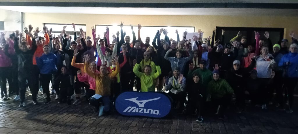Eine große Gruppe von Läufern in sportlicher Kleidung, die an einem frühen Morgen mit erhobenen Händen vor einem Gebäude posiert. Im Vordergrund steht ein Banner der Marke Mizuno.