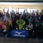 Eine große Gruppe von Läufern in sportlicher Kleidung, die an einem frühen Morgen mit erhobenen Händen vor einem Gebäude posiert. Im Vordergrund steht ein Banner der Marke Mizuno.