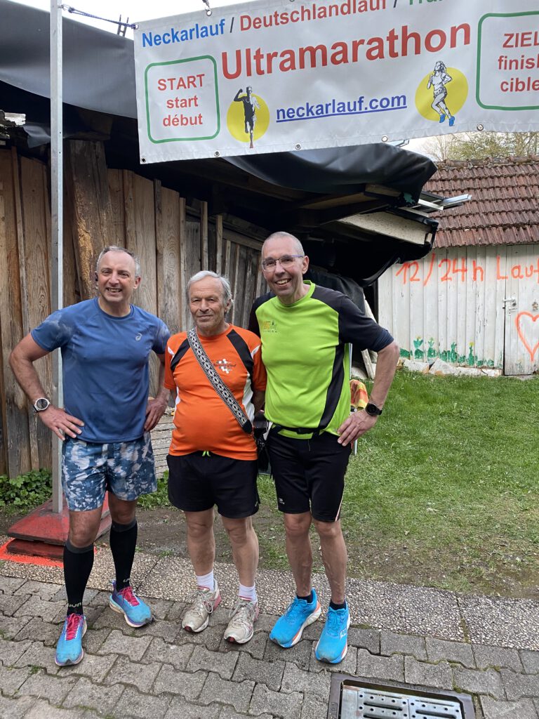Drei Männer stehen an einem Startpunkt für einen Ultramarathon. Der erste trägt ein blaues T-Shirt und schwarze Shorts, der zweite ein orangefarbenes T-Shirt mit einer Umhängetasche, und der dritte ist in einem grünen T-Shirt mit schwarzen Shorts gekleidet. Im Hintergrund ist das Banner des Events sichtbar.