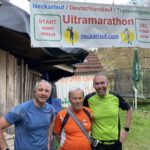 Drei Männer stehen lächelnd vor einem Banner mit der Aufschrift "Ultramarathon". Das Banner zeigt die Begriffe "START" und "ZIEL" sowie die URL neckarlauf.com. Im Hintergrund sind eine Holzhütte und eine grüne Liege sichtbar.