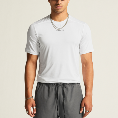 Craft Essence Tee Männer - Sportshirt in Weiß, aus atmungsaktivem Material, abgebildet auf einem Mann, der auch eine graue Sportshorts und eine weiße Kappe trägt, während er Sonnenbrillen trägt.