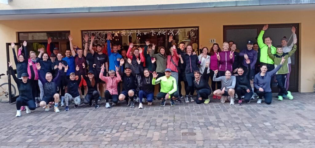 Eine große Gruppe von Läufern steht vor einem Gebäude und hebt die Hände in die Luft. Die Teilnehmer tragen sportliche Kleidung in verschiedenen Farben, darunter Schwarz, Pink und Gelb. Das Bild zeigt eine freundliche und einladende Atmosphäre.