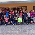 Eine große Gruppe von Läufern steht vor einem Gebäude und hebt die Hände in die Luft. Die Teilnehmer tragen sportliche Kleidung in verschiedenen Farben, darunter Schwarz, Pink und Gelb. Das Bild zeigt eine freundliche und einladende Atmosphäre.