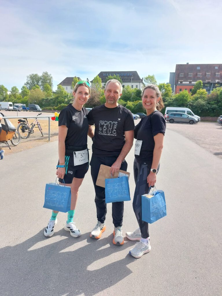 Drei Personen stehen auf einem Parkplatz nach einem Lauf. Zwei Frauen tragen schwarze T-Shirts und halten blaue Taschen. Ein Mann in der Mitte trägt ebenfalls ein schwarzes T-Shirt und hält eine gebräunte Urkunde. Im Hintergrund sind parkende Fahrzeuge und grüne Bäume zu sehen.