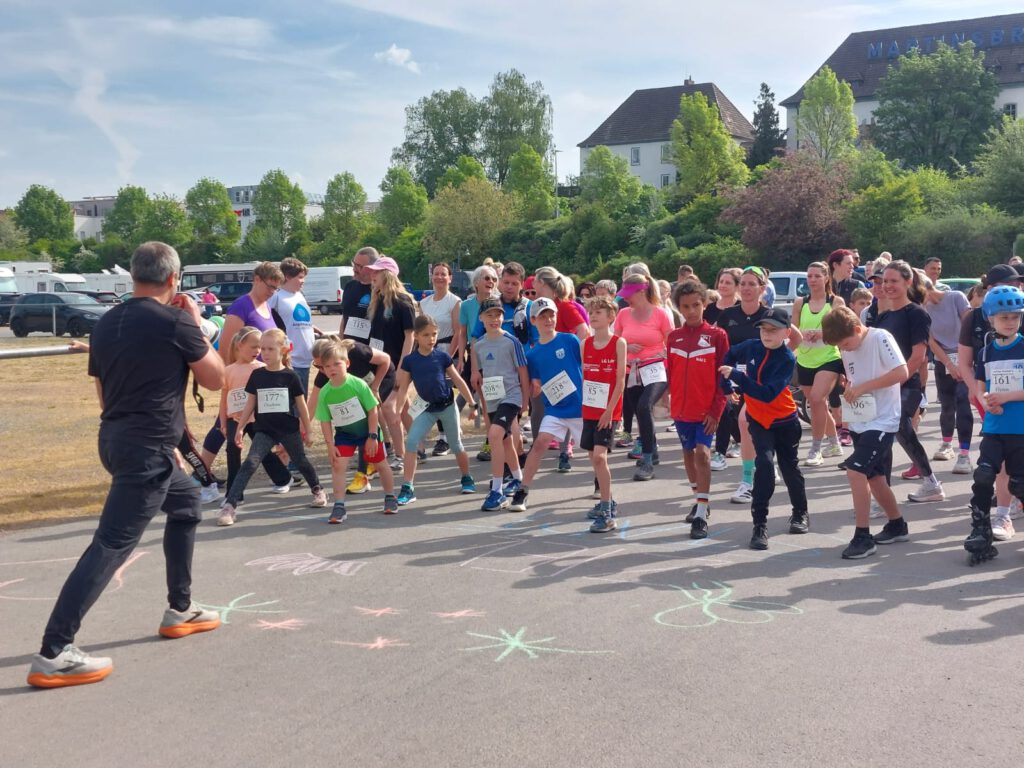 Eine Gruppe von Kindern in bunten T-Shirts startet zu einem Laufwettbewerb. Im Vordergrund steht ein Erwachsener, der sie anfeuert. Im Hintergrund sind Bäume und Gebäude sichtbar sowie einige Zuschauer.