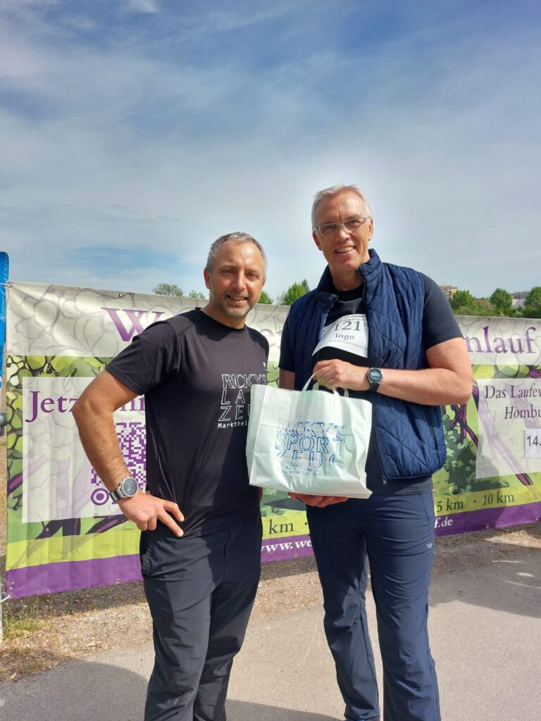Zwei Männer stehen nebeneinander vor einem Hintergrund mit einem Banner. Der Mann links trägt ein schwarzes T-Shirt und hält eine Tasche, während der Mann rechts ein blaues Oberteil und eine Startnummer trägt. Beide lächeln.