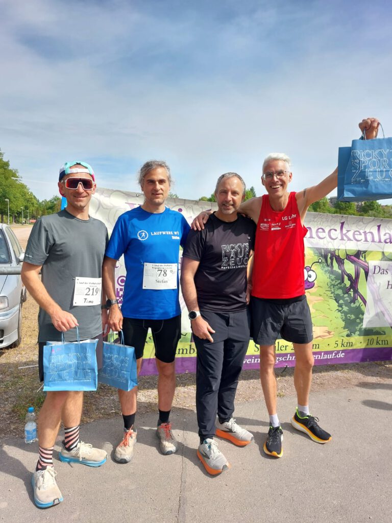 Vier Männer stehen nach einem Lauf zusammen, tragen Sportkleidung und halten die Geschenke in den Händen. Sie stehen vor einem bunten Banner der Veranstaltungsstelle, das den Lauf und die Distanz beschreibt. Die Umgebung ist hell und freundlich.