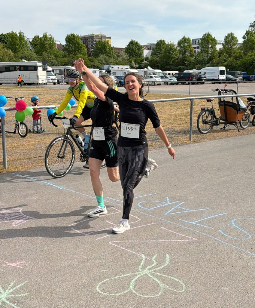 Zwei Läuferinnen, die beim Start eines Rennens fröhlich die Hände heben. Im Hintergrund sind Radfahrer und bunte Ballons zu sehen. Auf dem Boden sind bunte Kreidezeichnungen und die Aufschrift "START" zu erkennen.