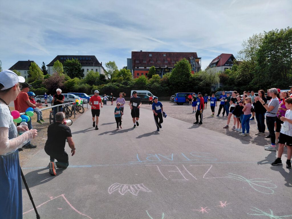 Eine Gruppe von fünf Läufern, gekleidet in Sportbekleidung, sprintet auf einer Straße. Zuschauer stehen am Rand, manche halten Luftballons. Im Hintergrund sind mehrere Gebäude und parkende Autos sichtbar. Der Himmel ist klar und blau.
