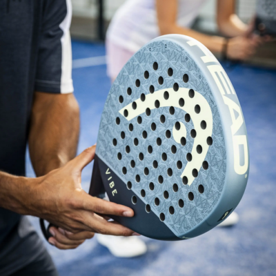 Padel Schläger Vibe von HEAD in blauer Farbe, mit einem charakteristischen Design und perforierter Oberfläche, gezeigt in der Hand eines Spielers auf dem Platz.