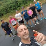 Eine Gruppe von acht Joggern steht auf einer regennassen Straße. Im Vordergrund zeigt ein Mann mit einem schwarzen T-Shirt und einer Trinkflasche in der Hand den Daumen nach oben. Die Gruppe trägt sportliche Kleidung und lächelt ins Kamera.