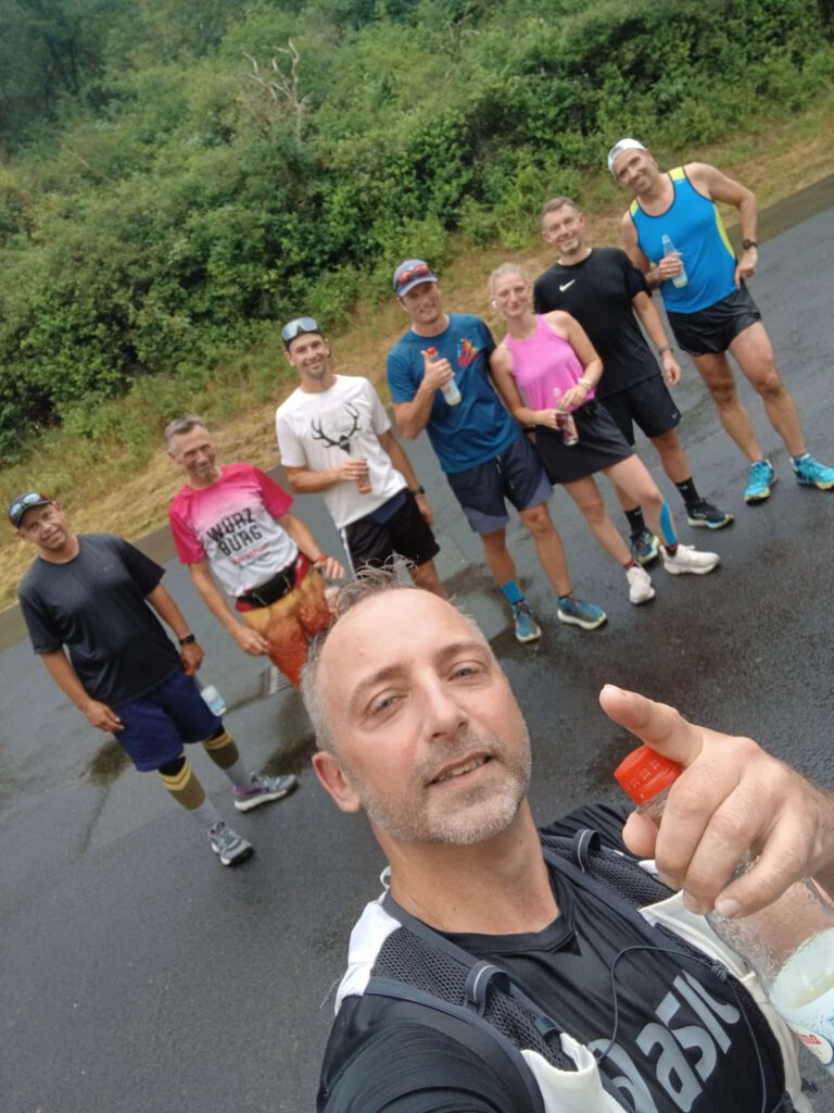 Eine Gruppe von acht Joggern steht auf einer regennassen Straße. Im Vordergrund zeigt ein Mann mit einem schwarzen T-Shirt und einer Trinkflasche in der Hand den Daumen nach oben. Die Gruppe trägt sportliche Kleidung und lächelt ins Kamera.