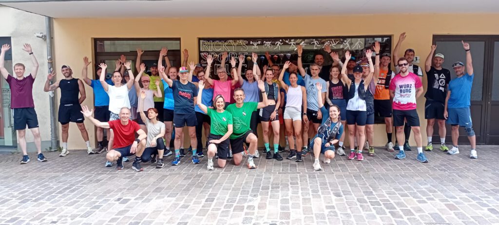 Eine Gruppe von über 30 Läufern steht vor einem Gebäude und hebt die Arme in die Höhe. Sie tragen Sportkleidung in verschiedenen Farben und posieren zusammen auf einem gepflasterten Platz.