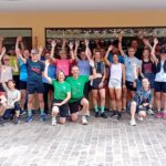 Eine Gruppe von über 30 Läufern steht vor einem Gebäude und hebt die Arme in die Höhe. Sie tragen Sportkleidung in verschiedenen Farben und posieren zusammen auf einem gepflasterten Platz.
