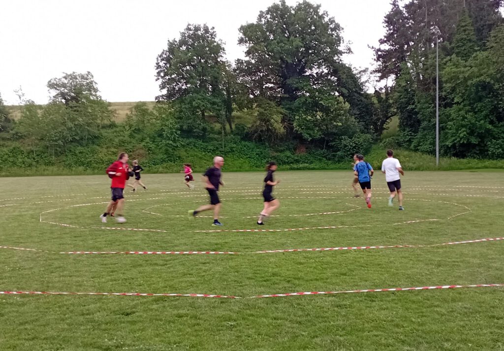 Eine Gruppe von sechs Personen joggt auf einem Rasenplatz, umgeben von Bäumen. Der Boden ist mit roten und weißen Markierungen in Kreisform abgetrennt, während der Himmel bewölkt ist.