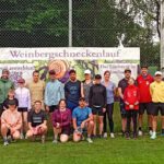 Eine Gruppe von Sportlern steht in Reihen auf einem Fußballplatz vor einem Banner mit der Aufschrift "Weinbergschneckenlauf". Im Hintergrund sind ein Fußballtor und Bäume zu sehen, und die Teilnehmer tragen Sportbekleidung.