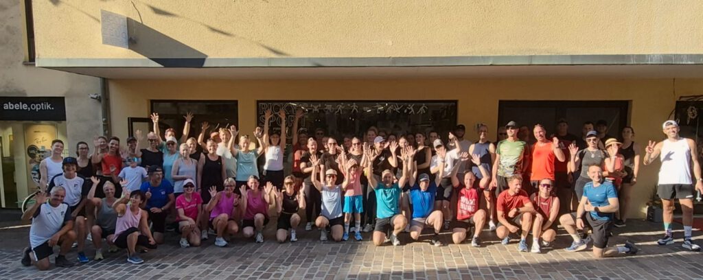 A large group of runners posing outside a building with their arms raised, standing on a cobblestone street under a yellow facade.