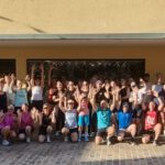 A large group of runners posing outside a building with their arms raised, standing on a cobblestone street under a yellow facade.