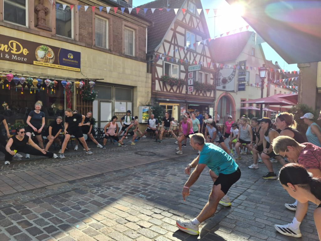 Runners stretch in a cobblestone street during a group event, with half-timbered buildings, shopfronts, and pennant flags lining the background.