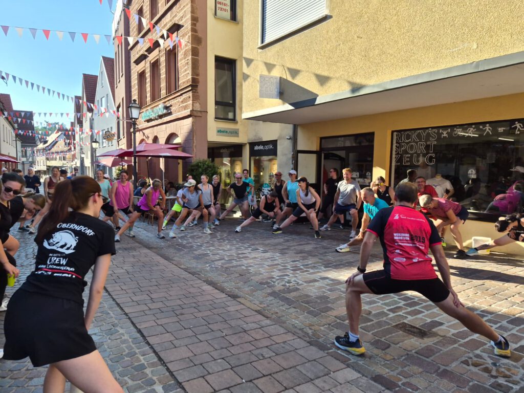 A group of runners stretches on a cobblestone street between buildings, with colorful bunting strung overhead and a crew member in a black shirt in the foreground.