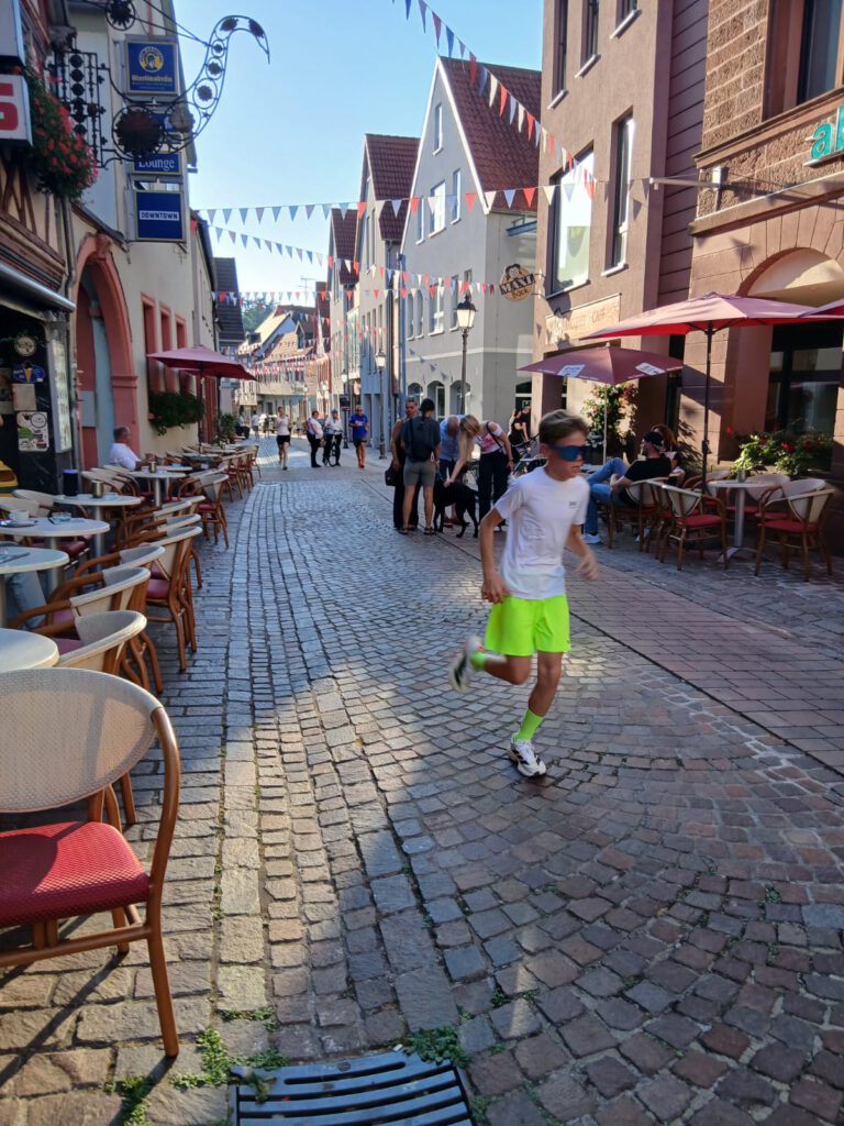 A runner in a white shirt and neon green shorts passes along a cobblestone street lined with café tables, umbrellas, and hanging bunting.